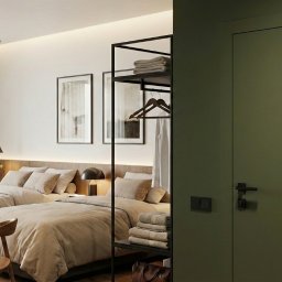 RICHTER Architekci - Nowoczesna sypialnia z dwoma łóżkami, stolikiem z laptopem i minimalistycznym regałem na ręczniki, oświetlona ciepłym światłem, z zielonymi drzwiami w tle.