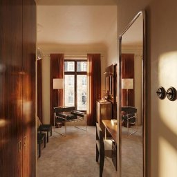 RICHTER Architekci - Eklektyczne wnętrze pokoju z biurkiem, krzesłem i szafą z ciemnego drewna, widziane przez lustro. Okno z widokiem na budynek w tle. Ciepłe oświetlenie.