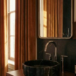 RICHTER Architekci - Stylowa łazienka z umywalką z czarnego marmuru, chromowaną armaturą i zasłonami w odcieniach brązu. Lustro z zaokrąglonymi rogami odbija wnętrze.