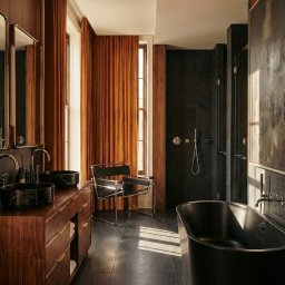 RICHTER Architekci - Elegancka łazienka z drewnianą szafką pod umywalki z marmurowymi misami, czarną wanną i kabiną prysznicową. Okno z pomarańczowymi zasłonami i krzesło.