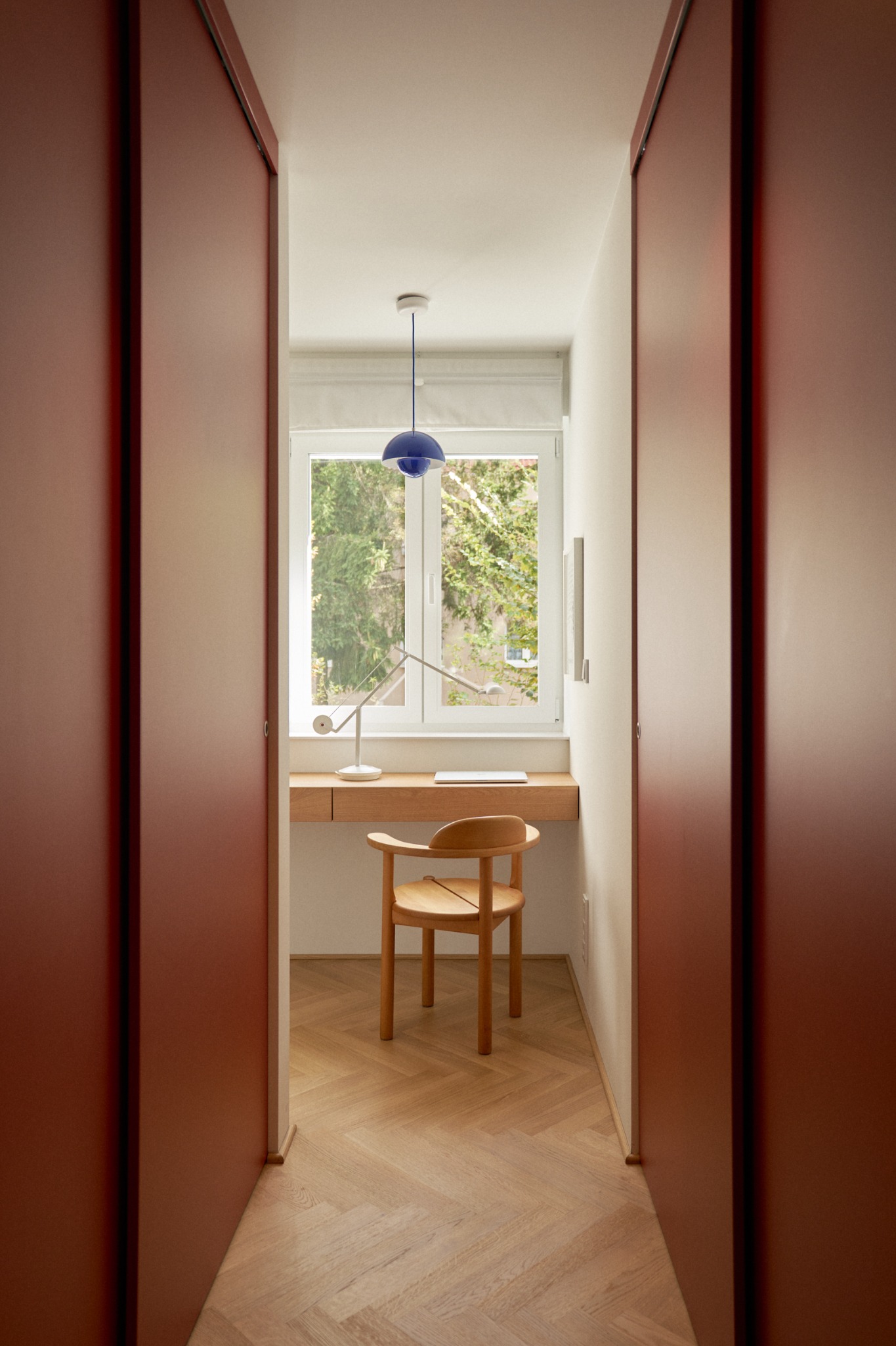 Wąski pokój z biurkiem pod oknem, drewnianym krzesłem, niebieską lampą wiszącą i podłogą w jodełkę, widziany zza rozsuwanych drzwi w kolorze terakoty.