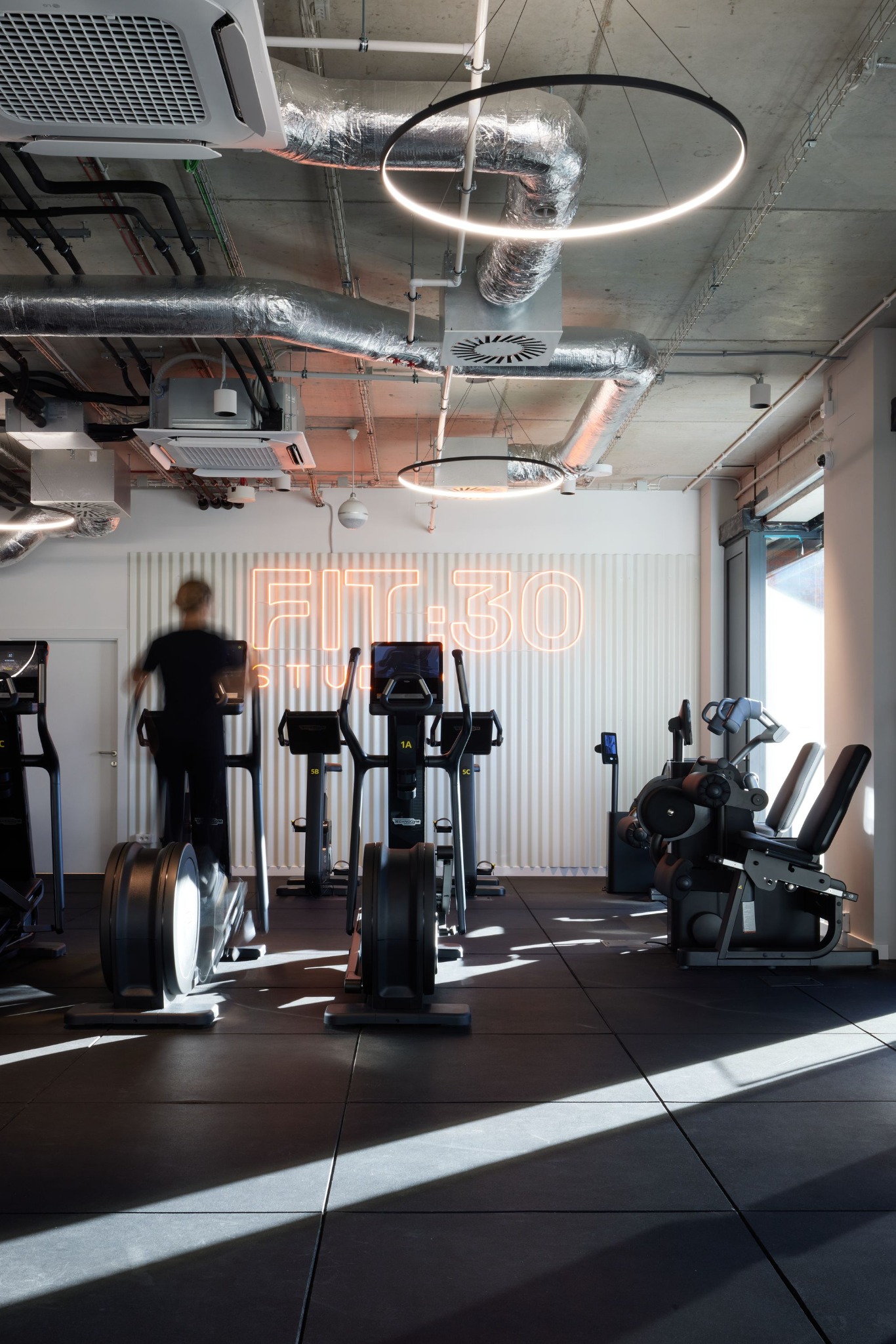 Nowoczesna siłownia z neonowym napisem 'FIT 30 STUDIO' na ścianie, widoczne maszyny do ćwiczeń i osoba korzystająca z bieżni. Oświetlenie sufitowe w kształcie okręgów.