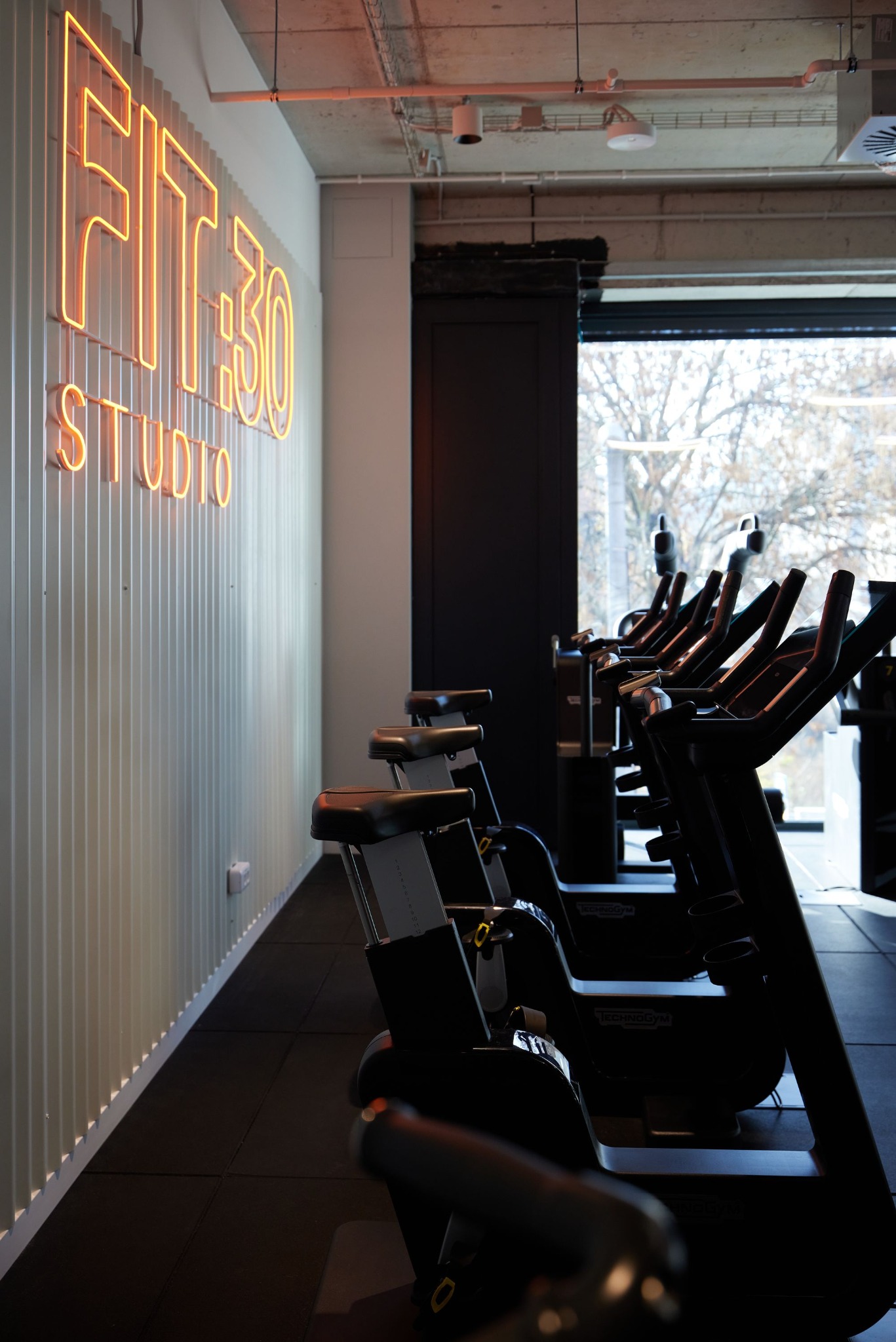 Nowoczesne studio fitness z rzędem czarnych rowerów stacjonarnych Technogym, neonowym napisem 'FIT30 STUDIO' na białej, żebrowanej ścianie i widokiem na drzewa za oknem.