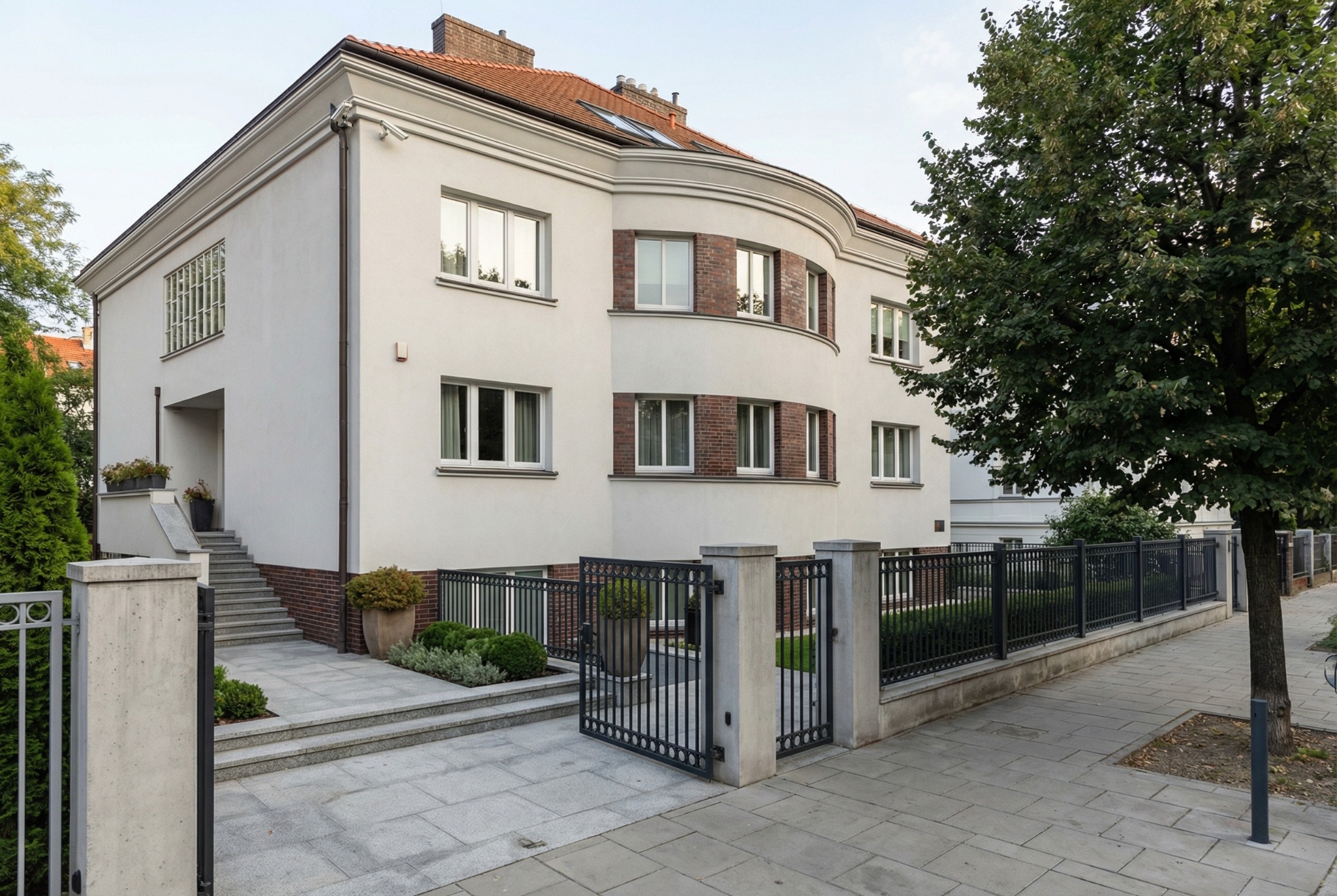Elegancki, dwukondygnacyjny dom z płaskim dachem i elementami z cegły klinkierowej. Widoczna brama wjazdowa i zadbany ogród. Jasna elewacja i duże okna.