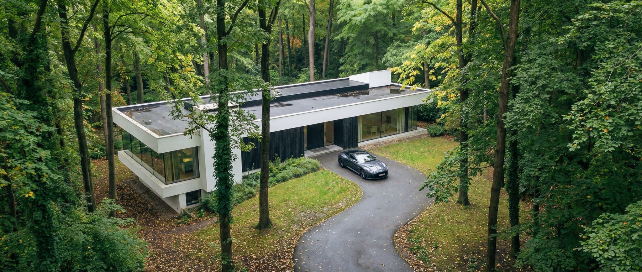 Nowoczesny dom z płaskim dachem, otoczony lasem, z szarym sportowym samochodem na podjeździe. Minimalistyczna architektura wkomponowana w naturalne otoczenie.