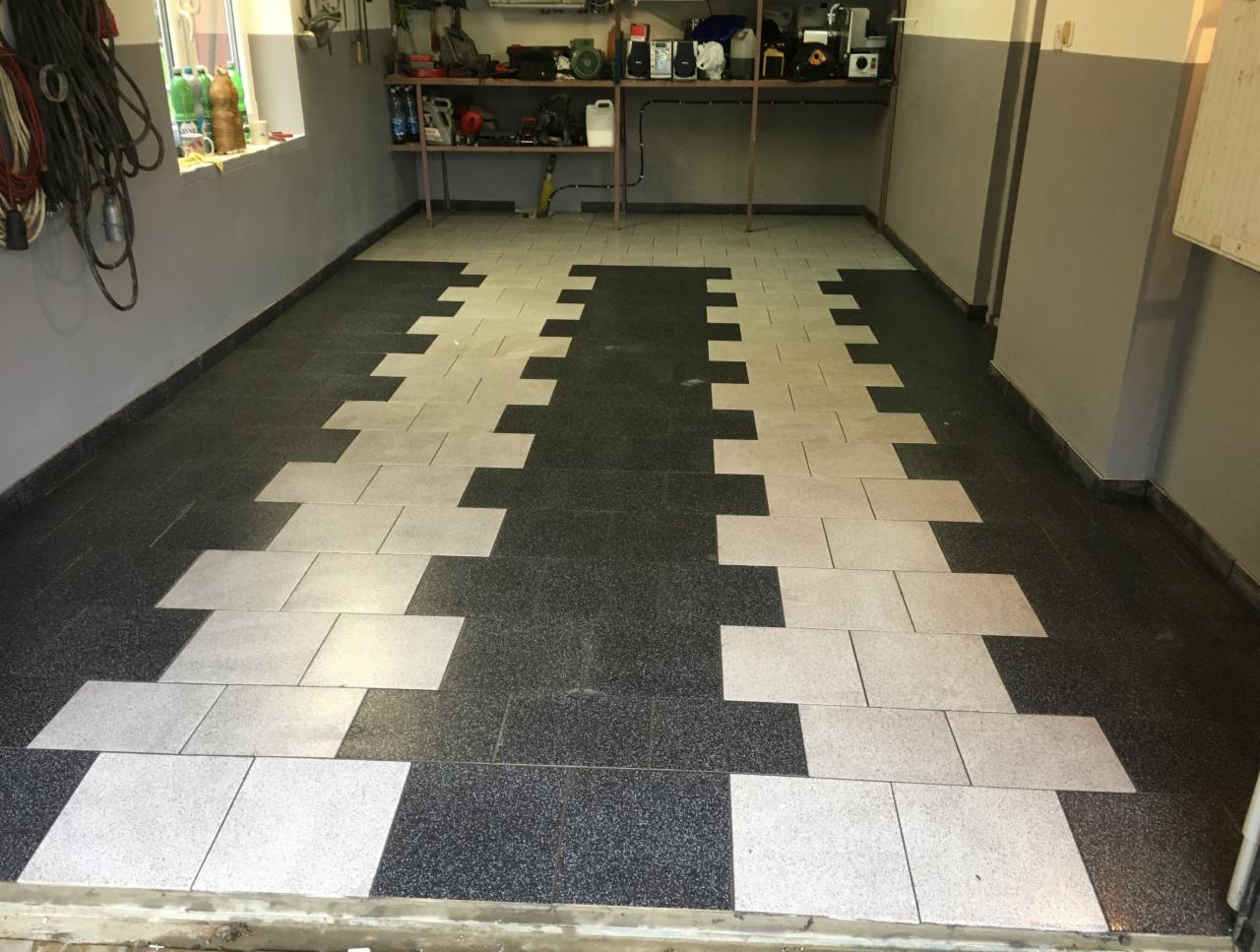 Układanie jasnych płytek na ciemnej posadzce w garażu, tworzących wzór przypominający ścieżkę, widoczne narzędzia na półce w tle.
