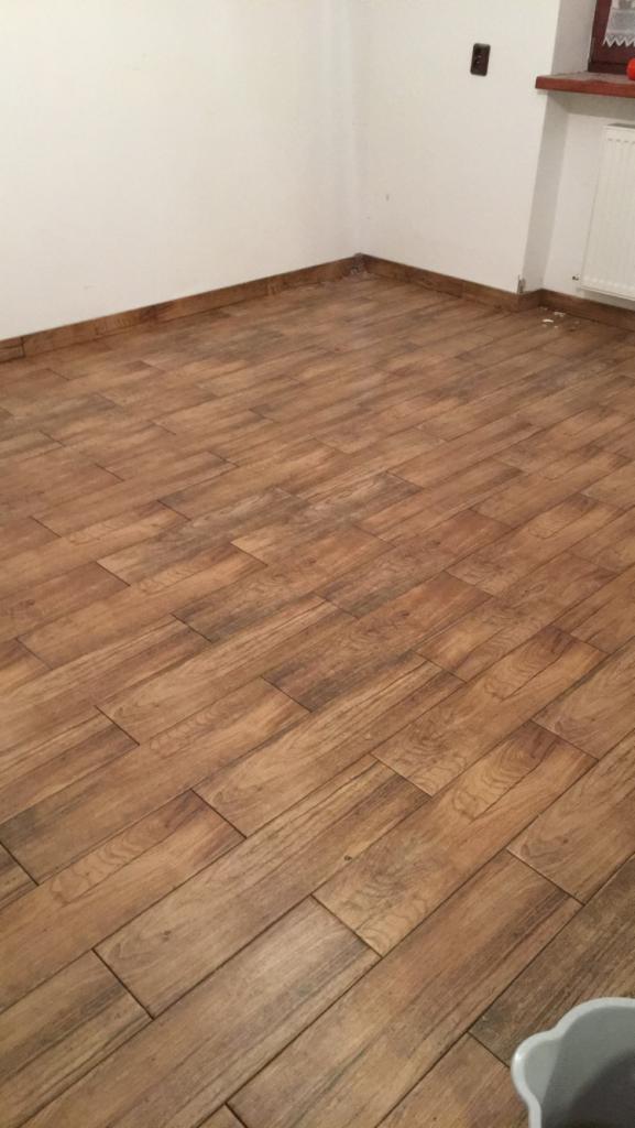 Ułożona podłoga z brązowych płytek imitujących drewno, widoczna ściana z listwą przypodłogową, fragment grzejnika i gniazdko elektryczne.