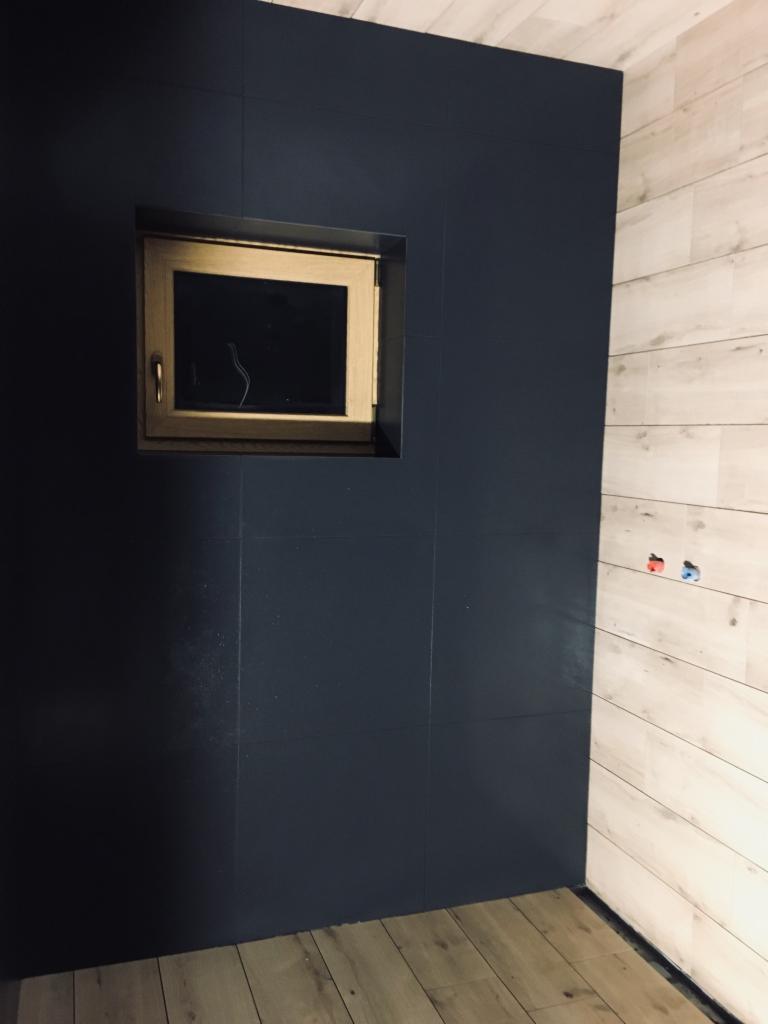Ciemnogranatowe płytki ścienne z małym okienkiem w łazience w Starym Sączu, kontrastujące z jasnymi panelami drewnopodobnymi na sąsiedniej ścianie i drewnianą podłogą.