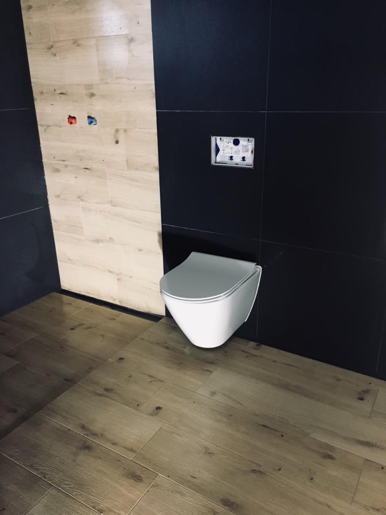 Nowoczesna toaleta podwieszana na tle czarnych i beżowych płytek imitujących drewno, z widocznymi przyłączami wodnymi na ścianie.