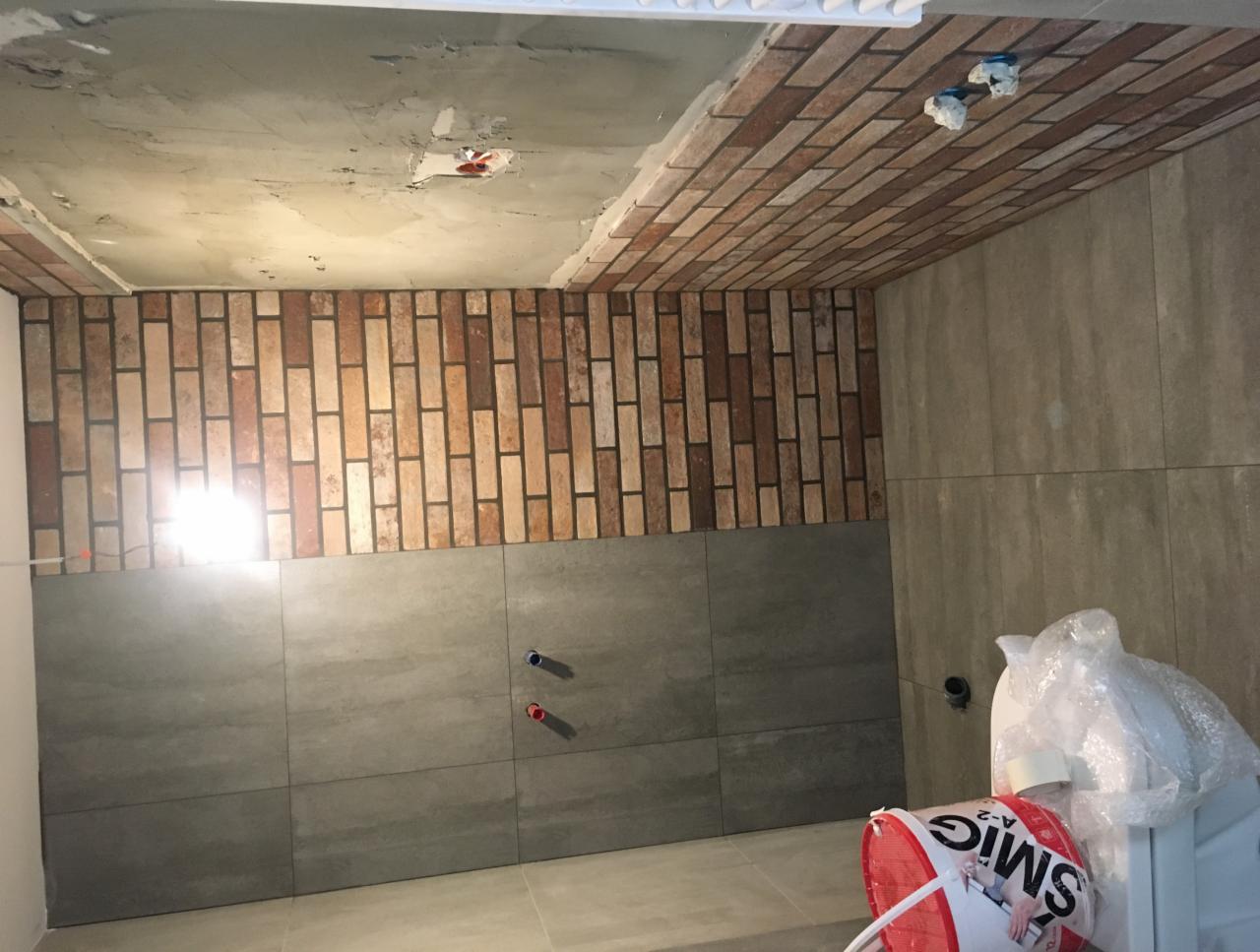 Wnętrze łazienki w trakcie remontu: częściowo ułożona ceglana glazura na ścianie i suficie, szare płytki, widoczne instalacje, kubeł z klejem i zabezpieczona folią toaleta.