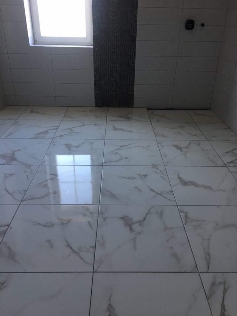 Wykończona łazienka z białymi płytkami podłogowymi imitującymi marmur i szarym pionowym pasem dekoracyjnym na ścianie, widoczne białe fugi i okno w górnym lewym rogu.