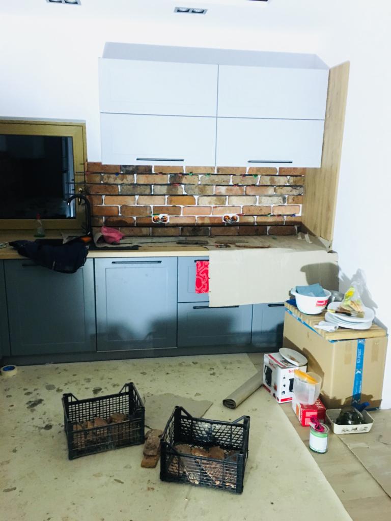 Fragment kuchni w trakcie prac wykończeniowych: widoczne szare szafki, białe szafki wiszące, ściana wyłożona cegłą z widocznymi miejscami na gniazdka elektryczne, kartony i pojemniki z akcesoriami...