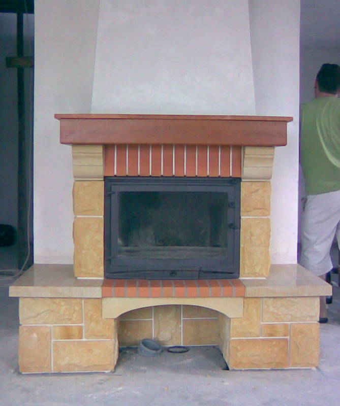 Murowany kominek z wkładem, obłożony kamieniem i cegłą, z drewnianym blatem, w trakcie remontu, widoczne niedoskonałości wykończenia.