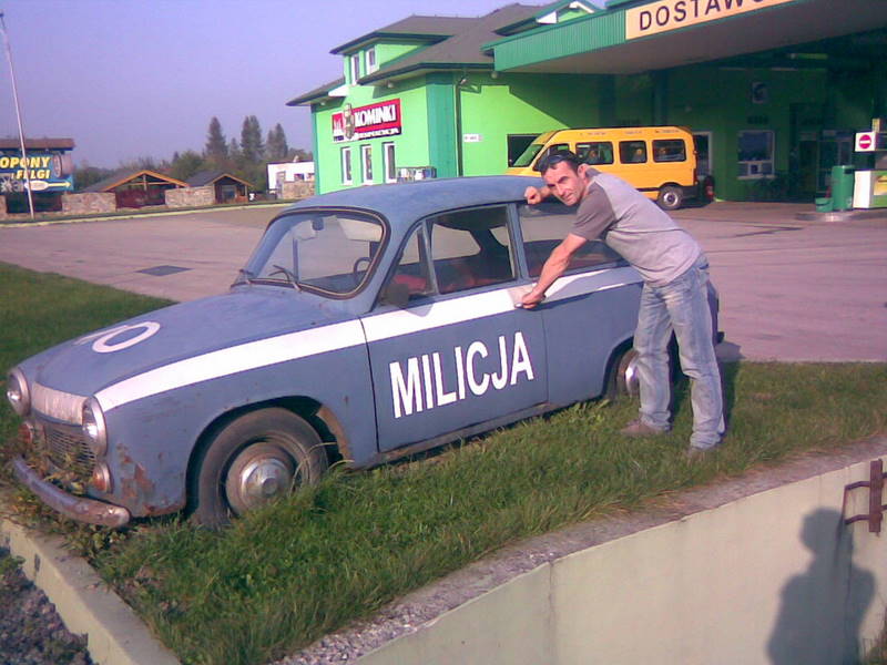 Mężczyzna opiera się o zardzewiały, niebieski samochód milicyjny z napisem 'MILICJA', stojący na trawie przed budynkiem z zielonym dachem i żółtym busem w tle.