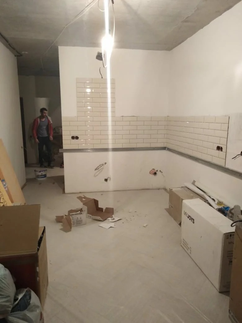 Wnętrze kuchni w trakcie układania białych, prostokątnych płytek na ścianie; widoczne puszki elektryczne, kartony na podłodze i osoba w tle.