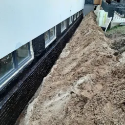 Odkopane fundamenty budynku z nałożoną hydroizolacją, widoczny grunt i fragment budynku z oknami.