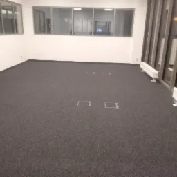 Wnętrze biurowe z ciemnoszarą wykładziną dywanową, widoczne kratki podłogowe, białe grzejniki pod oknami i duże okna z pionowymi żaluzjami.