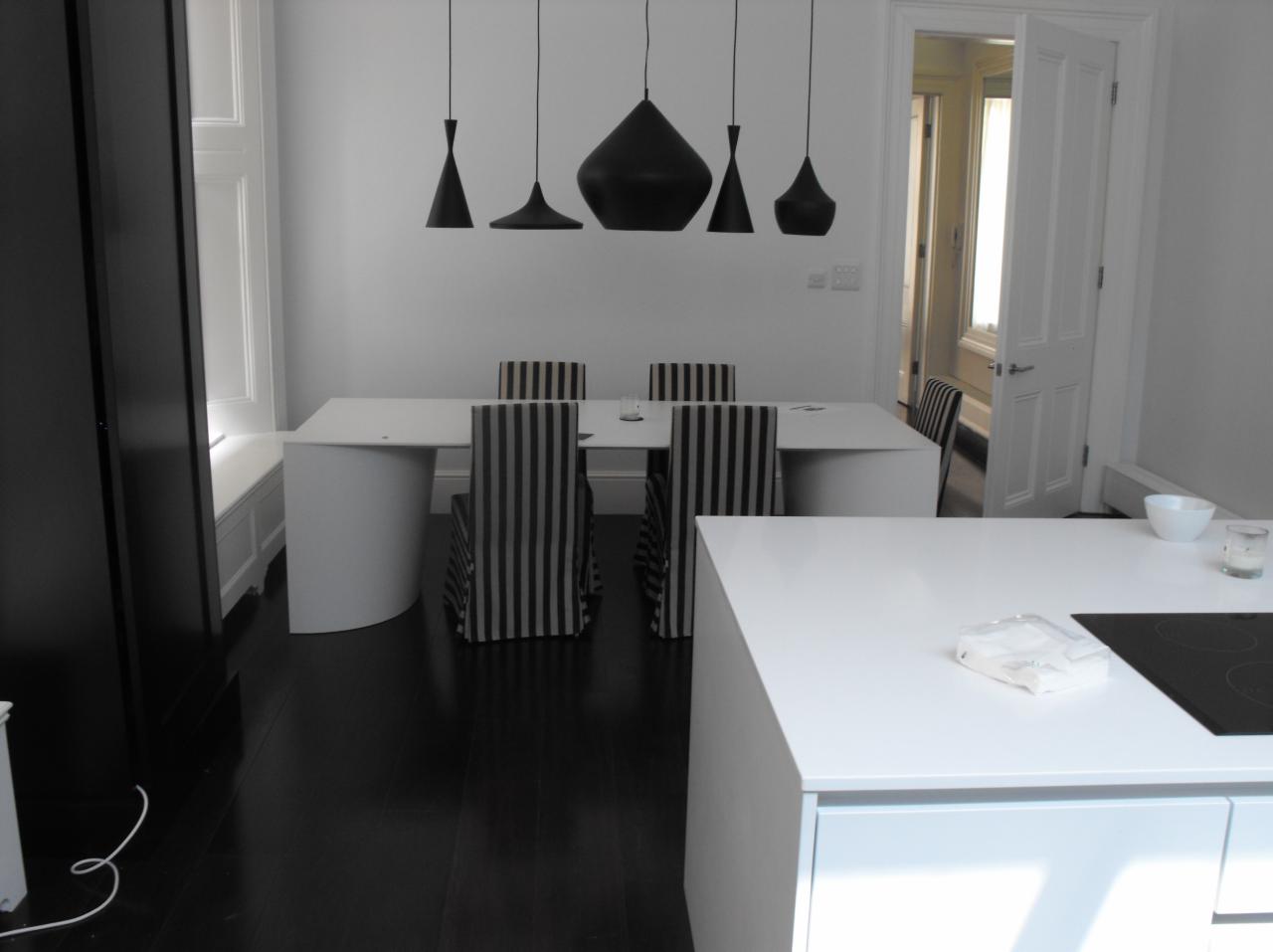Minimalistyczna jadalnia z czarnymi lampami wiszącymi nad białym stołem i krzesłami w czarno-białe pasy, ciemna podłoga, widok na otwarte drzwi do innego pomieszczenia.