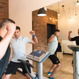 Dwóch mężczyzn podczas treningu EMS w studio, jeden w specjalnym kombinezonie z elektrodami, drugi w koszulce polo, obaj z uniesionymi rękami.