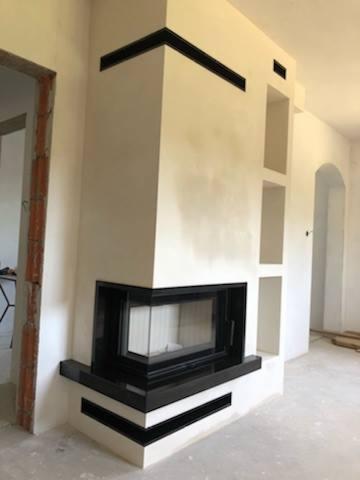 Nowoczesna obudowa wkładu kominkowego z czarnymi elementami dekoracyjnymi i półkami w ścianie, surowy stan wykończenia wnętrza.