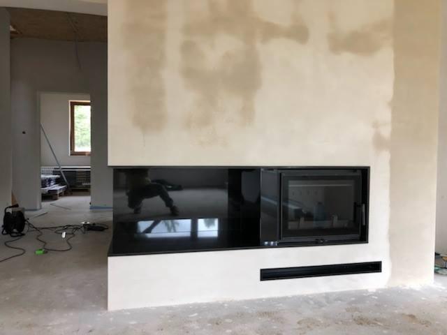 Nowoczesny wkład kominkowy z czarnym szkłem, osadzony w surowej ścianie z widocznymi śladami tynku, odbijający sylwetkę osoby w szklanej powierzchni.