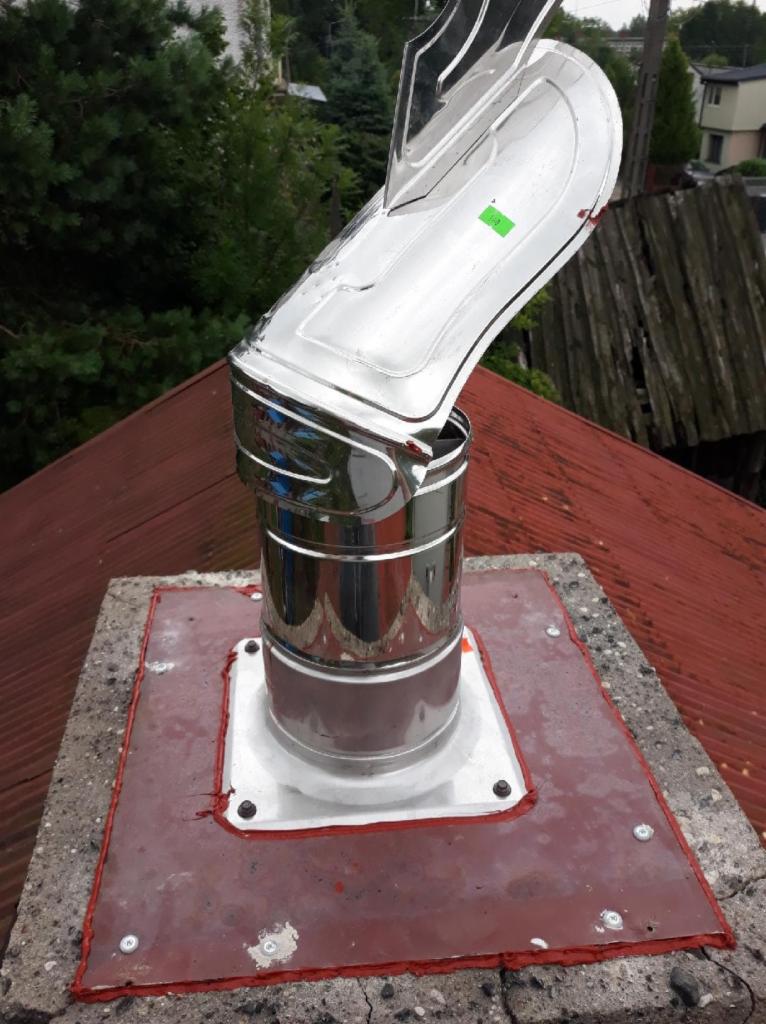 Błyszczący, chromowany wkład kominowy z daszkiem, zamontowany na betonowym kominie pokrytym czerwoną farbą, widok z góry na tle dachu i zieleni.