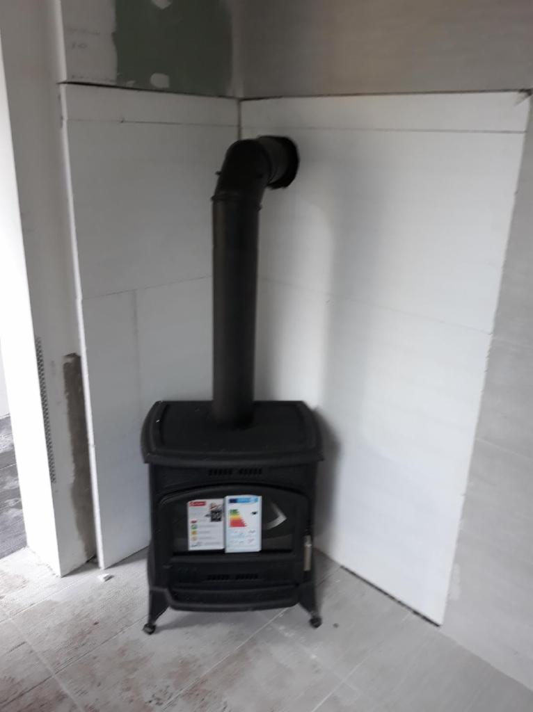 Czarny piec wolnostojący z rurą odprowadzającą dym, umieszczony w rogu pomieszczenia w trakcie wykańczania.