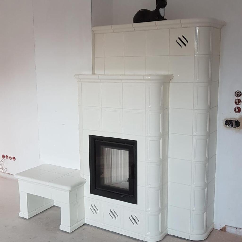 Biały piec kaflowy z czarnym wkładem kominkowym i dopasowanym taboretem, zwieńczony ceramiczną figurką kota, w trakcie montażu instalacji elektrycznej przy ścianie.