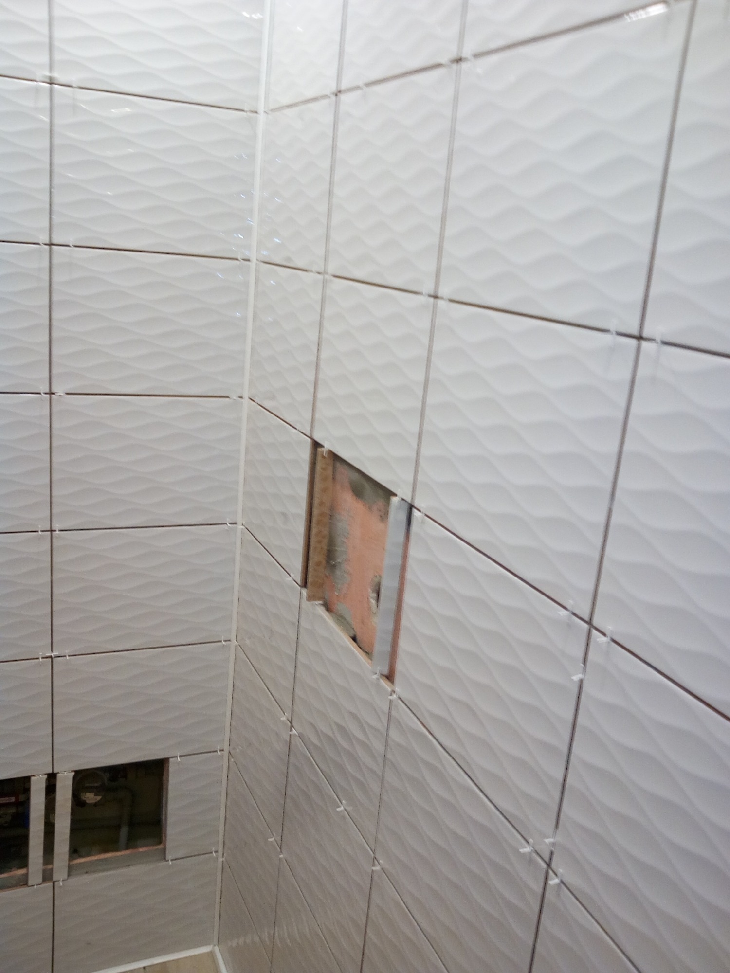 Ściana łazienki w trakcie układania białych płytek z falistym wzorem; widoczne krzyżyki dystansowe i otwory na instalacje.