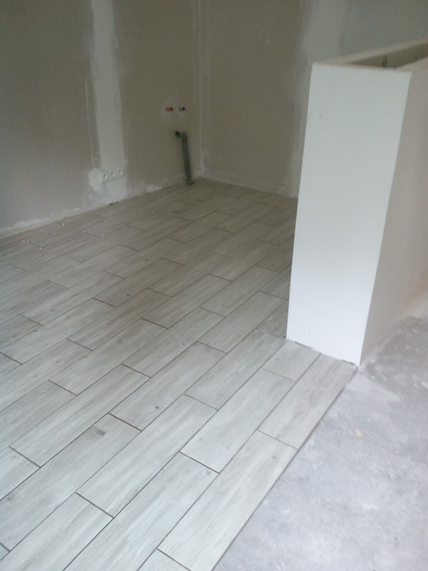 Ułożona, jasnoszara podłoga z płytek imitujących drewno w trakcie prac wykończeniowych. Widoczne surowe ściany i fragment zabudowy.