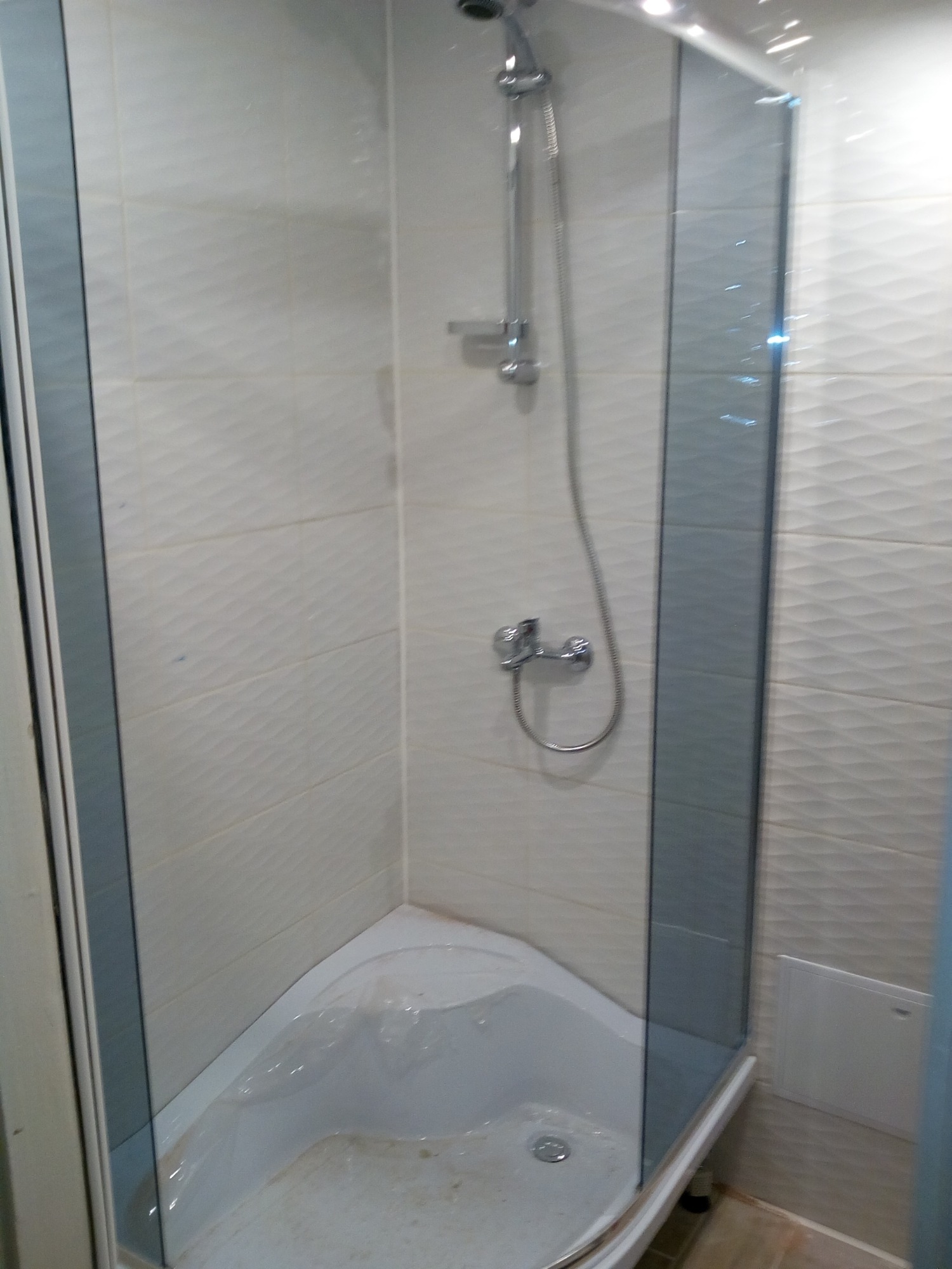 Narożna kabina prysznicowa z chromowaną armaturą, wykończona białymi płytkami z delikatną fakturą, zabezpieczona folią ochronną przed zabrudzeniami podczas prac wykończeniowych.