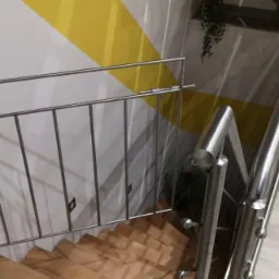 Metalowa barierka zabezpieczająca schody z drewnianymi stopniami, zamontowana na ścianie z żółtym akcentem kolorystycznym, widok z góry.