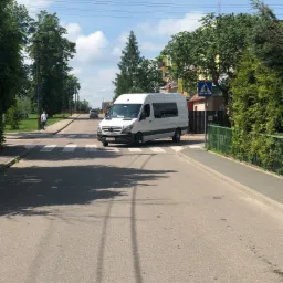 Biały bus marki Mercedes na przejściu dla pieszych w słoneczny dzień, widoczny pieszy po lewej stronie i budynki w tle.