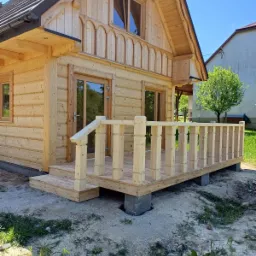 Jasny, drewniany dom z bali z werandą w trakcie budowy, widoczne betonowe bloczki fundamentowe i świeżo posiana trawa.