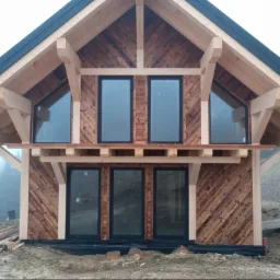 Surowy stan domu szkieletowego: drewniana konstrukcja z zamontowanymi oknami, ukośne deski elewacyjne, widoczny etap budowy z elementami rusztowania.