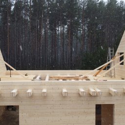 SimpleEnergy - Surowa konstrukcja domu z jasnego drewna na tle ciemnego lasu podczas opadów śniegu. Widoczne drabiny i narzędzia na budowie.