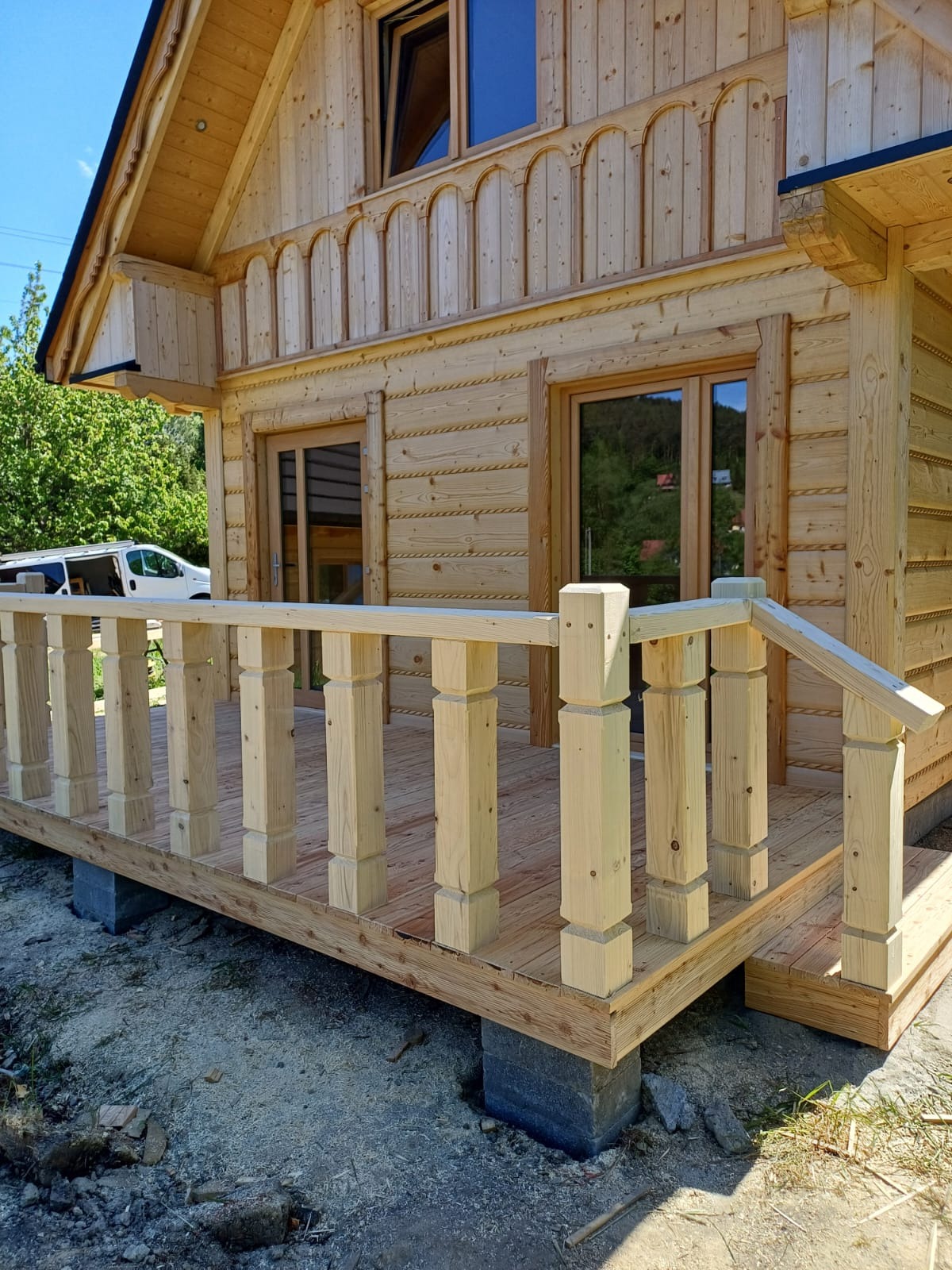 Surowy drewniany dom z bali w trakcie budowy, widoczny taras z balustradą i fragment elewacji z oknami, w tle częściowo widoczny samochód dostawczy.