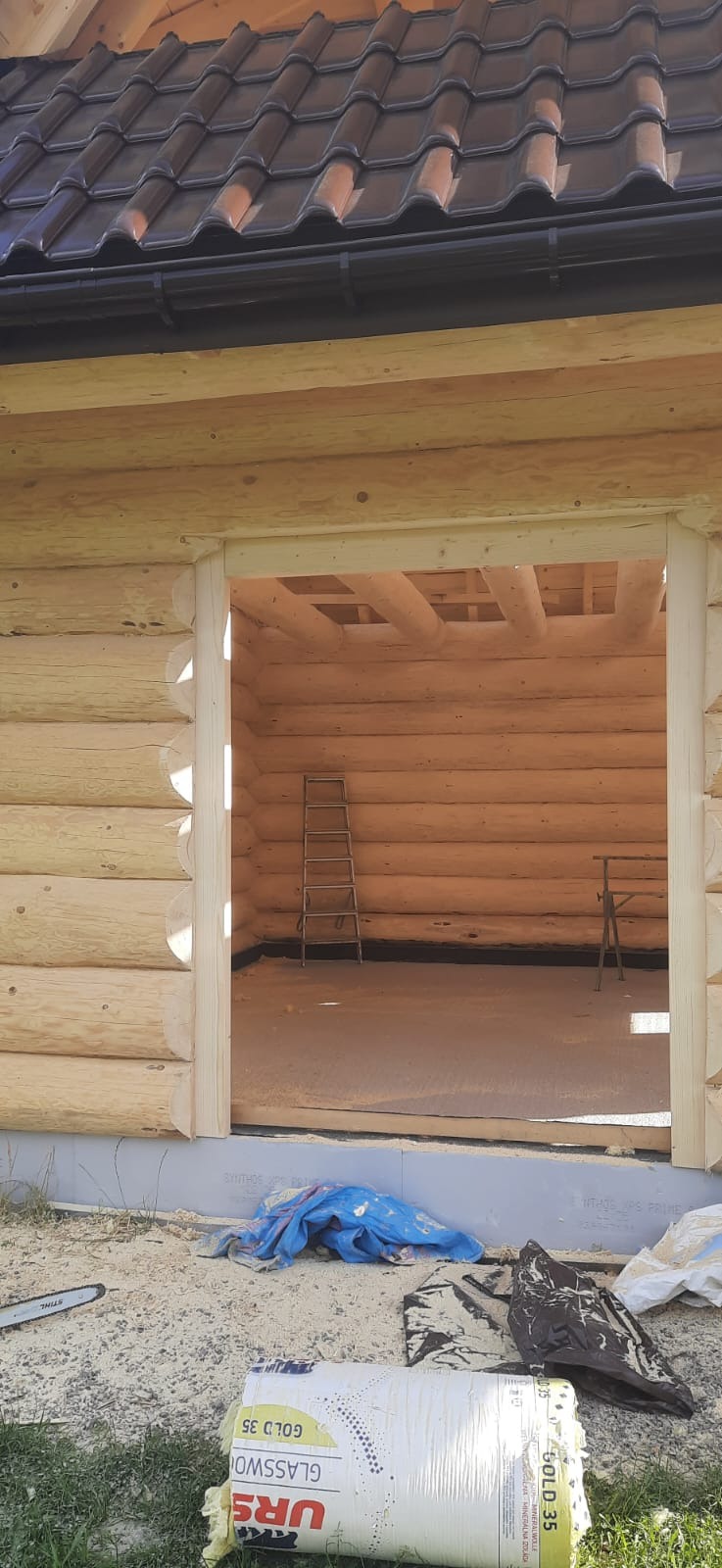 Wnętrze surowego domu z bali w trakcie budowy, widoczna drabina i stół roboczy w środku, na zewnątrz leży piła łańcuchowa i opakowanie wełny mineralnej.