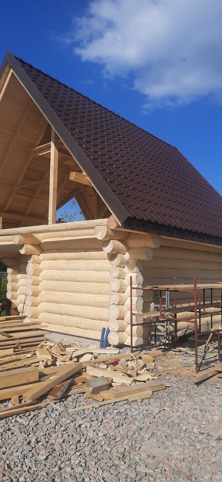 Surowy stan budowy domu z bali, widoczne bale z jasnego drewna, brązowy dach z metalowej dachówki, rusztowanie oraz nieuporządkowane deski i gruz na ziemi, błękitne niebo w tle.