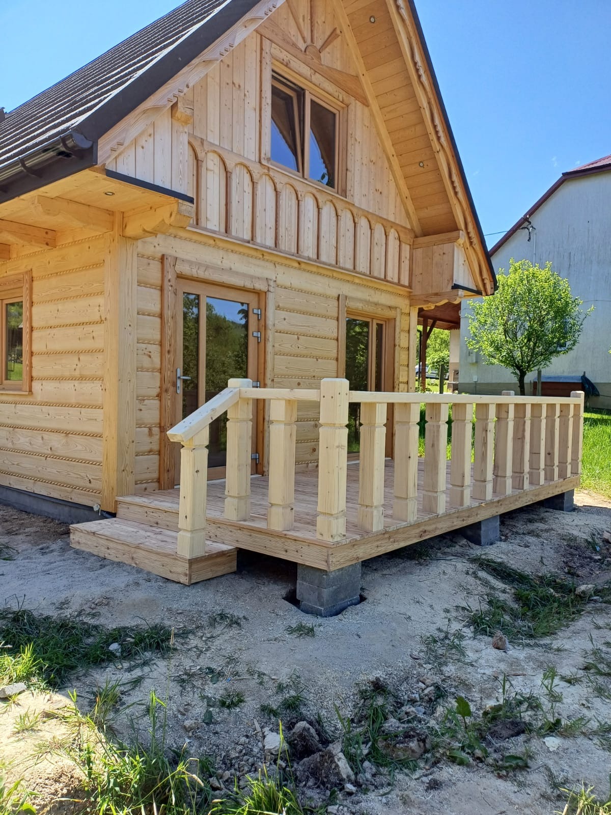 Jasny, drewniany dom z bali z werandą w trakcie budowy, widoczne betonowe bloczki fundamentowe i świeżo posiana trawa.