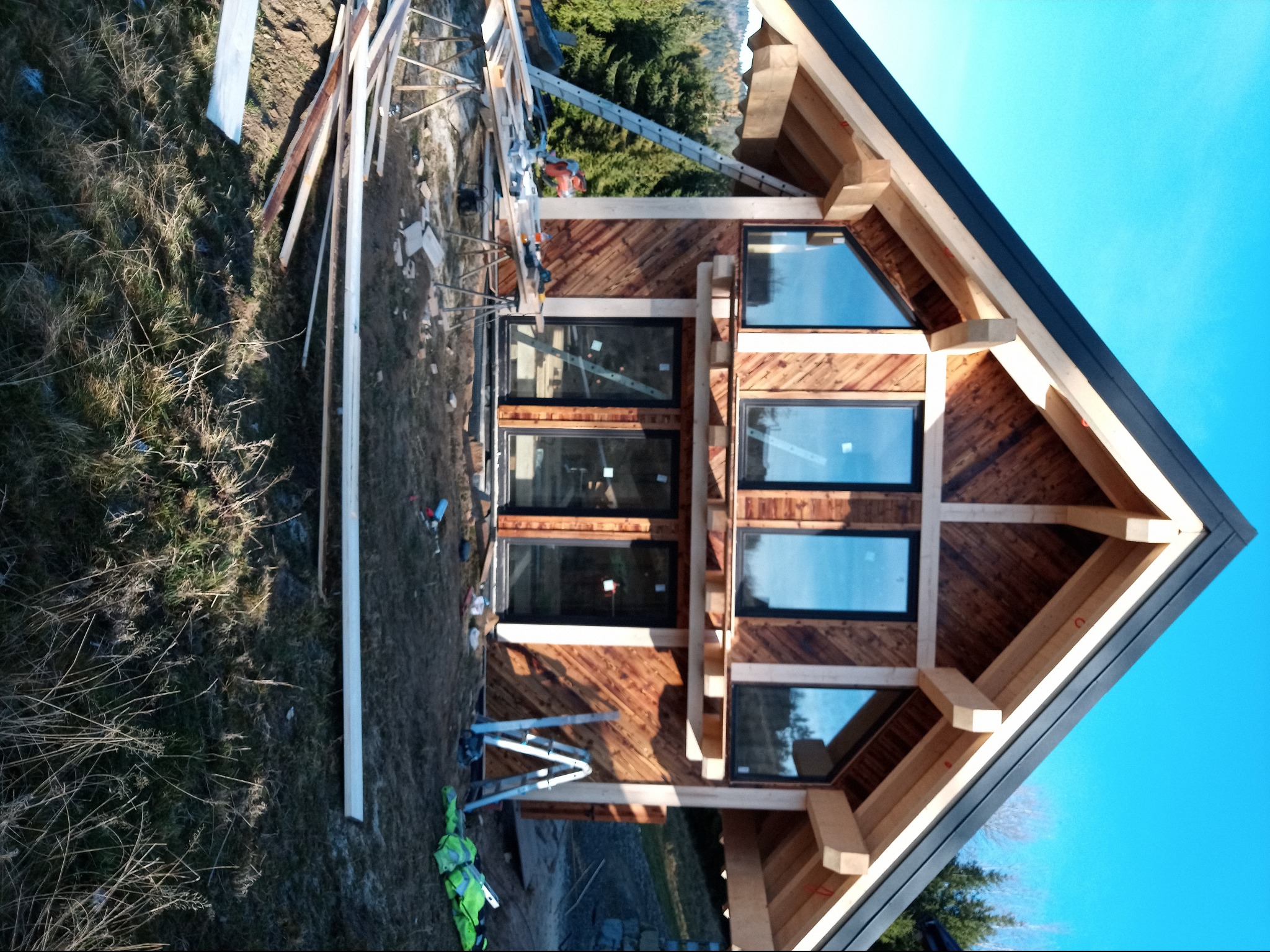 Wznoszenie drewnianego domu z dużymi oknami w czarnych ramach; widoczne elementy konstrukcyjne, rusztowania i teren budowy.