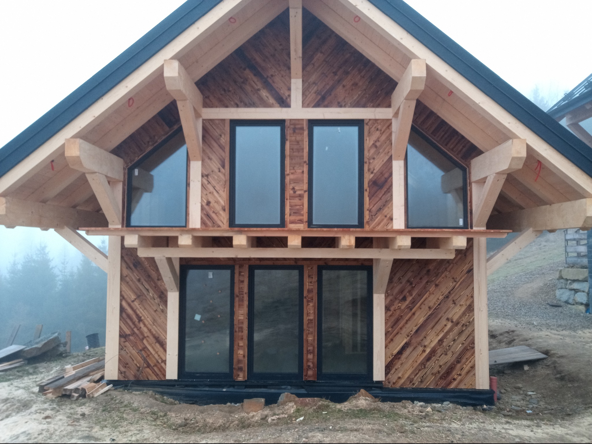 Surowy stan domu szkieletowego: drewniana konstrukcja z zamontowanymi oknami, ukośne deski elewacyjne, widoczny etap budowy z elementami rusztowania.