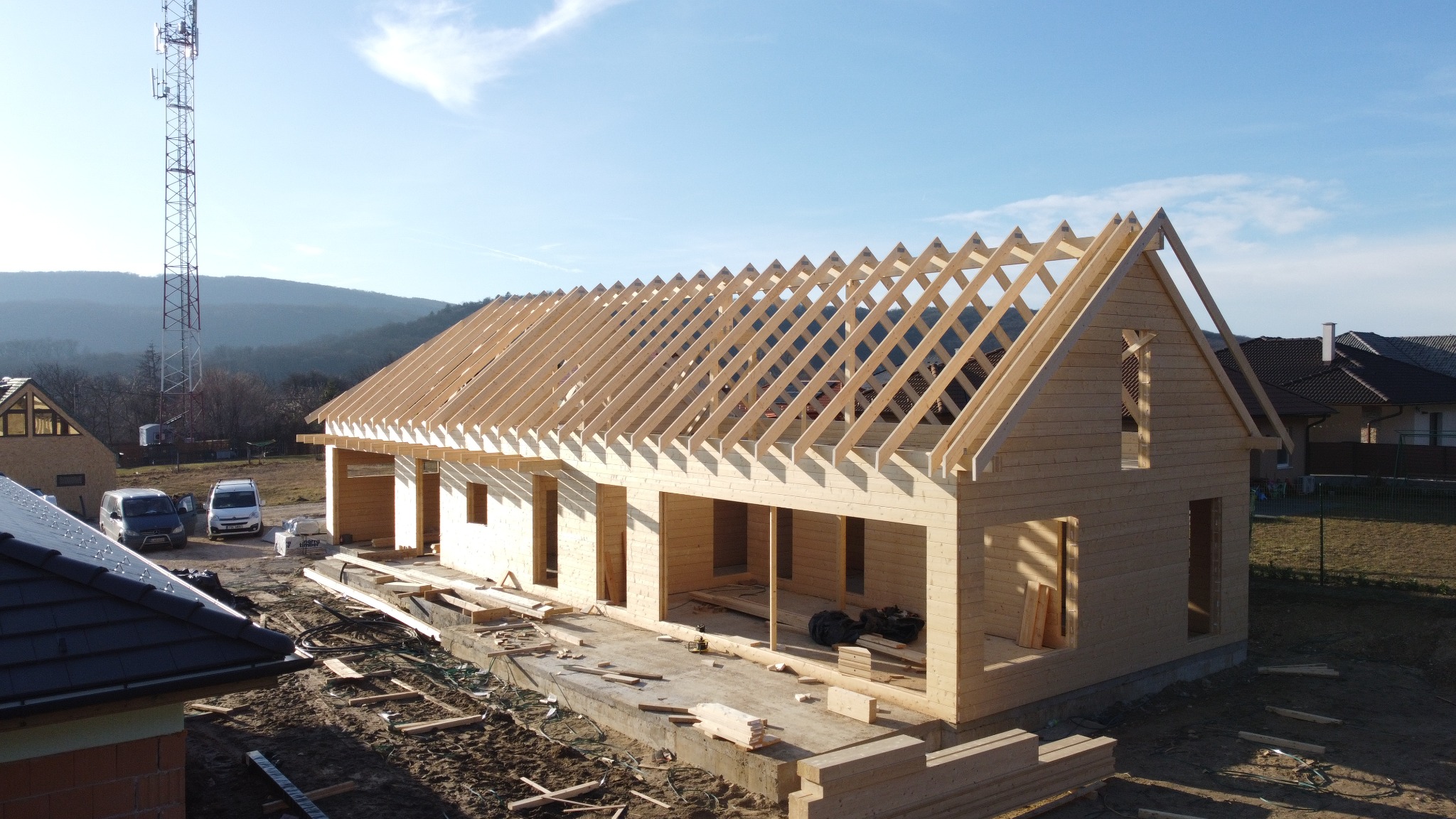 Wznoszenie domu z jasnego drewna, widoczne krokwie dachu i otwory okienne. W tle zabudowa jednorodzinna i maszt telekomunikacyjny.