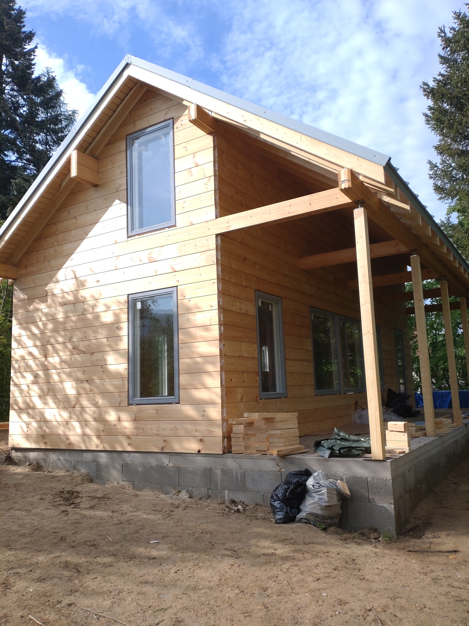 Surowy, drewniany dom w trakcie budowy z szarymi oknami i zadaszonym tarasem, na fundamencie z bloczków betonowych, otoczony zielenią.