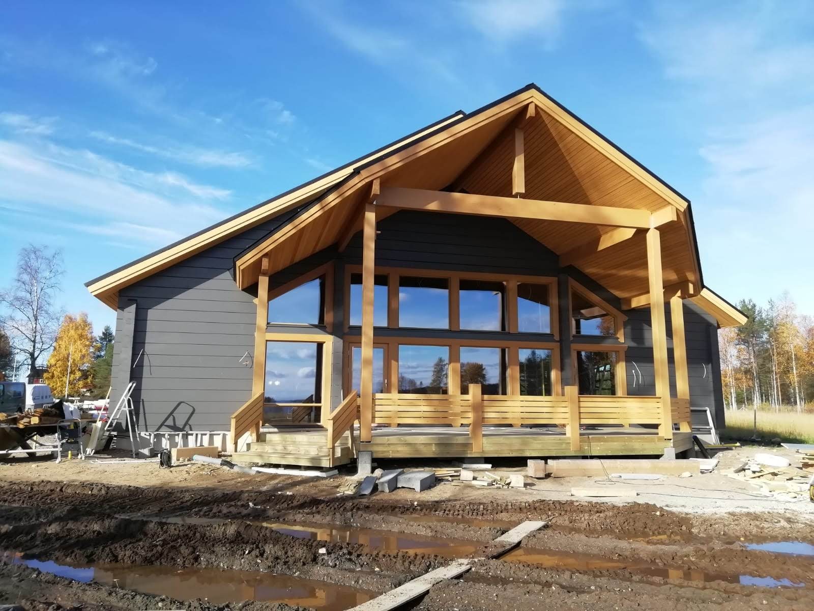 Surowy stan domu z bali z dużymi oknami i drewnianą konstrukcją, widoczne rusztowania i błoto na placu budowy, błękitne niebo w tle.