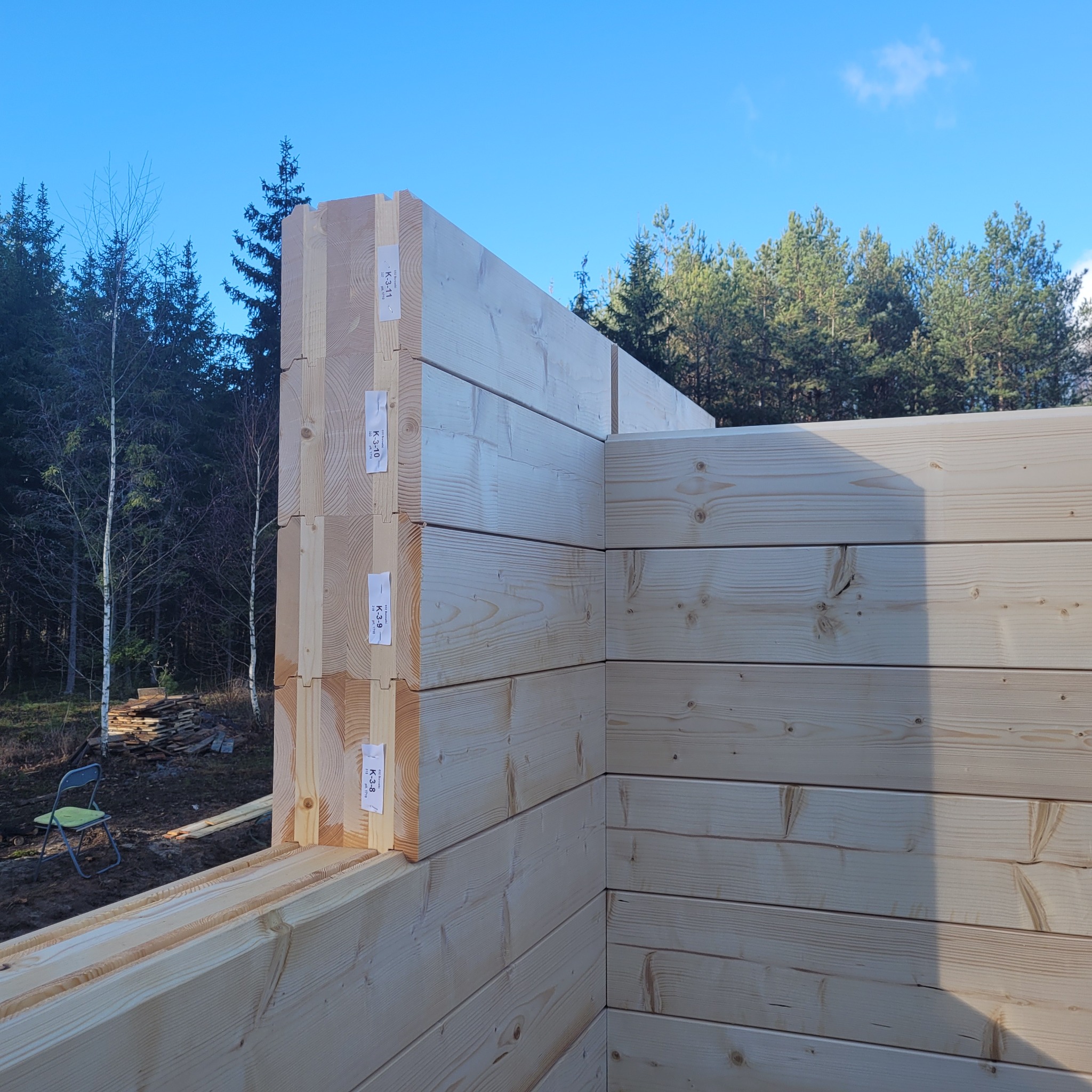Fragment ściany domu z bali w trakcie budowy, widoczne oznaczenia elementów, w tle las i błękitne niebo. Jasne drewno, precyzyjne łączenia.