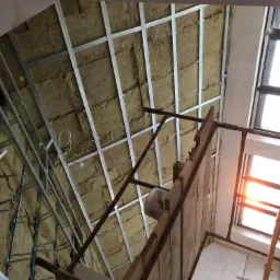 Skośny widok na poddasze w trakcie ocieplania: wełna mineralna między metalowymi profilami, widoczne rusztowanie z drewnianych desek i wiadro z klejem. W tle okno z jasnym światłem słonecznym.