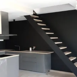 Nowoczesna kuchnia z szarymi szafkami, wyspą i czarną ścianą z drewnianymi schodami bez poręczy, widoczne narzędzia i pozostałości po pracach wykończeniowych.