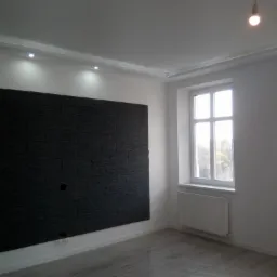 Pusty pokój po remoncie z czarną ścianą z imitacji kamienia, szarą podłogą, białymi ścianami i grzejnikiem pod oknem, z wiszącą żarówką bez klosza.