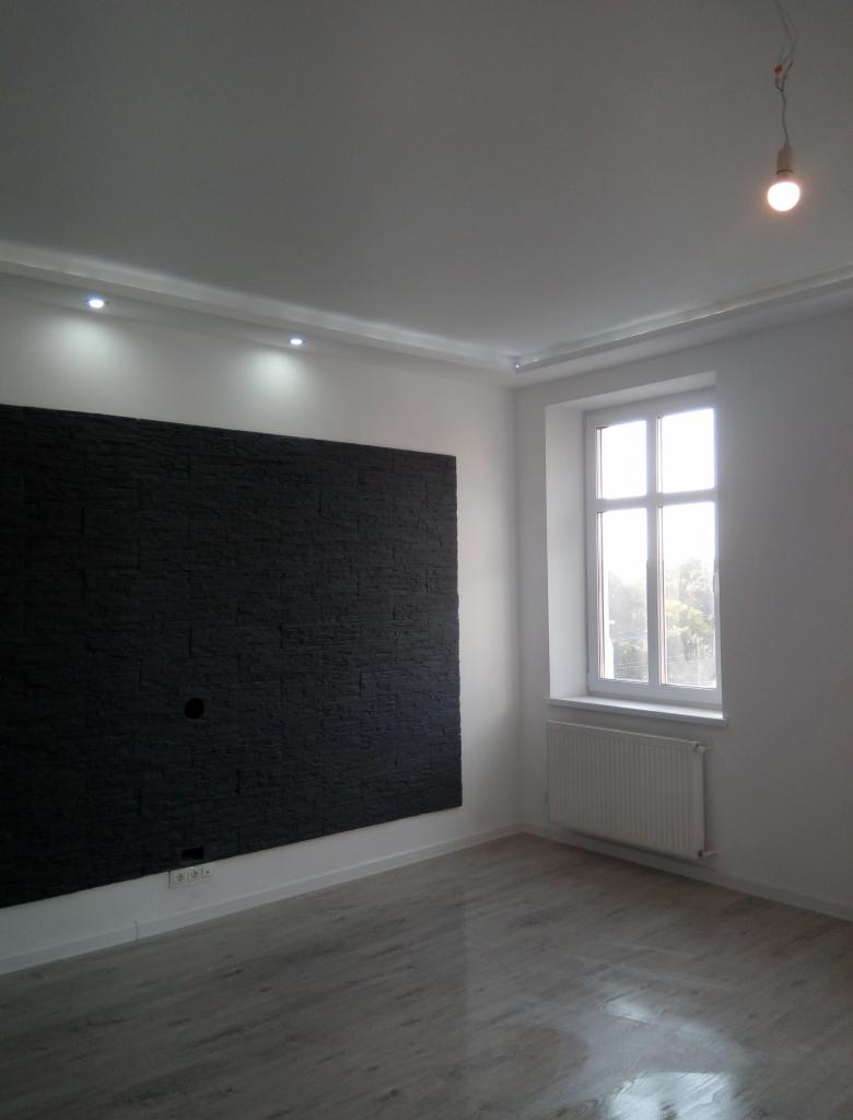Pusty pokój po remoncie z czarną ścianą z imitacji kamienia, szarą podłogą, białymi ścianami i grzejnikiem pod oknem, z wiszącą żarówką bez klosza.